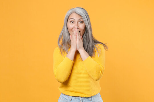 Shocked amazed surprised excited cheerful gray-haired asian woman wearing casual clothes standing covering mouth with hands looking camera isolated on bright yellow colour background, studio portrait.