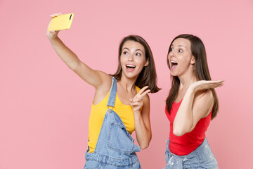 Two surprised funny young brunette women friends 20s wearing casual denim clothes doing selfie shot on mobile phone showing victory sign isolated on pastel pink colour background, studio portrait.