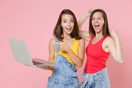 Two excited happy young brunette women friends 20s in casual denim clothes pointing index finger on laptop pc computer doing winner gesture isolated on pastel pink colour background, studio portrait.