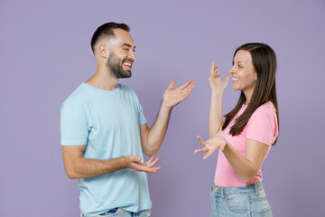 Smiling cheerful young couple two friends man woman wearing blue pink blank design t-shirts speaking talking spreading hands looking at each other isolated on pastel violet background studio portrait.