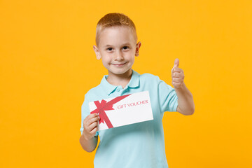 Smiling cute little fun male kid boy 5-6 years old wearing stylish blue turquoise t-shirt polo hold in hand mockups of gift certificate isolated on yellow color wall background, child studio portrait.