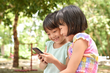 Two smiling little girl holding smartphone is watching online video in park outdoor.