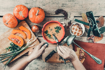 Pumpkin soup on wooden background, vintage. Woman hands, cooking process. Food blogger recording video with mobile phone on tripod, holding remote shutter. Thanksgiving, vegan. Flat lay, top view.