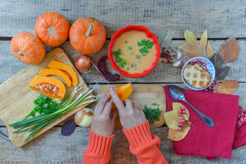 Pumpkin soup on wooden table with red linen cloth and vintage cutlery. Woman hands cutting vegetables, cooking process. Autumn vegetarian, healthy food concept. Thanksgiving dinner, top view.
