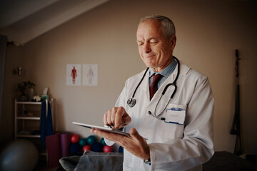 Standing senior doctor with stethoscope around neck holding digital tablet in hospital - happy Doctor using technology 