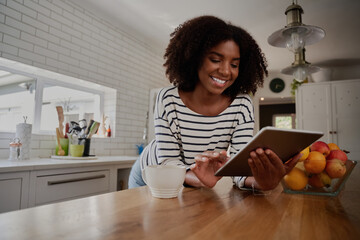 African American Woman standing in kitchen leaning on counter using digital tablet looking at online recipes 