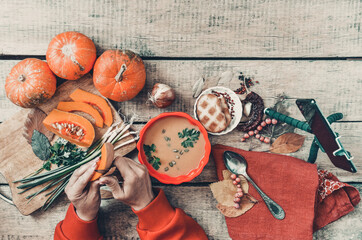 Pumpkin soup on wooden background, vintage. Woman hands, cooking process. Food blogger recording video with mobile phone on tripod, holding remote shutter. Thanksgiving, vegan. Flat lay, top view.