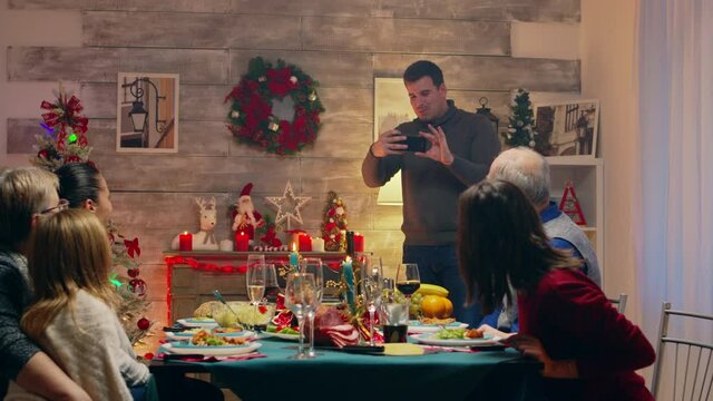 Cheerful man taking a photo of his beautiful family at christmas reunion. Traditional festive christmas dinner in multigenerational family. Enjoying xmas meal feast in decorated room. Big family