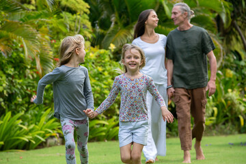 Young happy family walking in tropical park or gaden with palm trees. Two cute little sisters running in front of parents. Summer exotic vacation