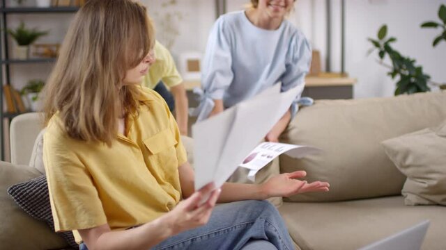 Handheld shot of annoyed mother sitting on couch in living room and looking through documents while working remotely, then getting frustrated as her misbehaved children bothering her