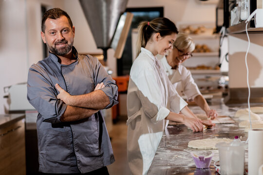 Portrait of mid adult male baker with coworkers in uniform preparing dough for baking bread in modern manufacturing.