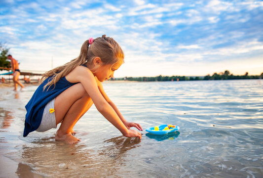 Side view of little lovely caucasian girl playing with tiny rubber yellow ducks in small blue pool, standing on beach sand. Attractive kid having fun, enjoying holidays at sea, summertime.
