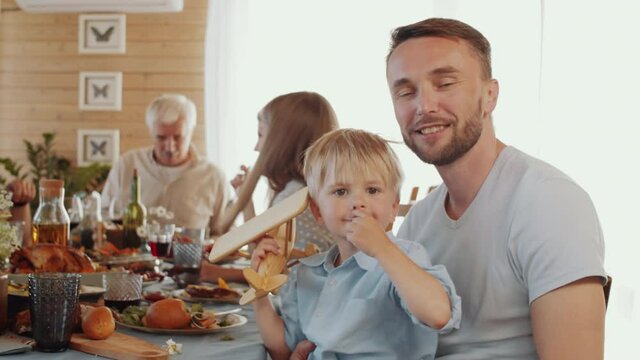 Handsome Caucasian man sitting with cute little son on his knees, looking at camera and smiling while having holiday dinner with family at home