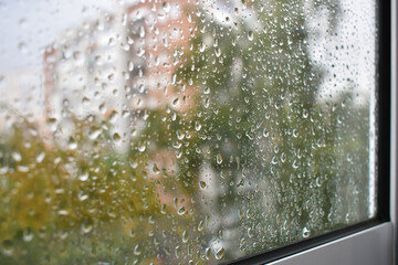 Raindrops on a rainy day on the window pane. Autumn cloudy weather through glass covered with rain drops. Blurred autumn cityscape. Selective focus on raindrops.