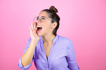 Young beautiful woman wearing glasses over isolated pink background shouting and screaming loud to side with hand on mouth