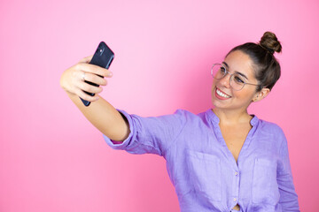 Young beautiful woman wearing glasses over isolated pink background taking a selfie with her phone