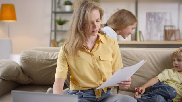 Medium shot of tired mother looking through documents and trying to work from home while her playful teenage girl and little boy jumping on couch and bothering her