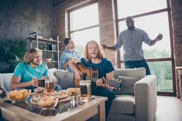 Four fellows enjoy celebrate college start holiday gather house hold pint mug indoors blonde hair man sit comfort couch play guitar afro american guy dance enjoy melody