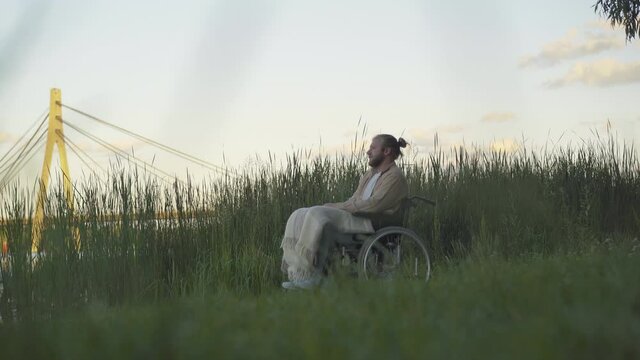 Wide shot of thoughtful young handicapped man on wheelchair sitting on river bank and admiring sunset. Side view portrait of handsome disabled Caucasian invalid enjoying summer evening outdoors.