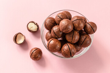 Raw macadamia nuts in a transparent glass bowl and scattered on pink solid background. Minimal composition.