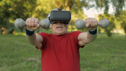 Senior old grandfather man in VR headset helmet making fitness exercises with dumbbells in park