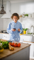 Happy Young Man with Long Hair Using Smartphone while Wearing Blue Pajamas. He is Scrolling News Feed, Social Media, Chatting and Drining Morning Coffe in a Kitchen with Healthy Vegetables.