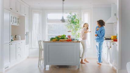 Handsome Young Man in Blue Pajamas and Beautiful Girl in Striped Jumper are Fooling Around in the Kitchen. White Modern Kitchen Area with Healthy Green Vegetables on a Table. Happy Couple in Cozy Home