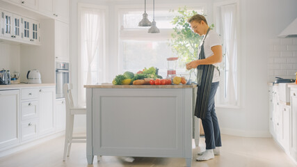 Hipster Young Man in Glasses Cooking on a Modern Kitchen. He Wears an Apron and Preparing a Vegetable Salad in a Sunny Bright Kitchen with Healthy Green Vegetables on a Table.