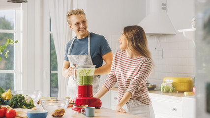 Handsome Young Man in Glasses Wearing Apron and Beautiful Girl are Making A Smoothie in the Kitchen. Happy Couple are Trying Healthy Organic Beverage. Male and Female at Home on a Sunny Day.