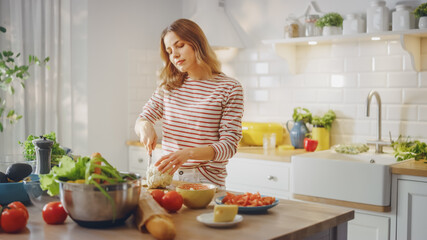 Young Female in Striped Jumper is Making a Healthy Organic Salad in a Modern Sunny Kitchen. Authentic Woman is Chopping a Cauliflower with a Knife. Natural Clean Diet and Healthy Way of Life Concept.