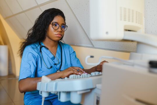 Young african-american female doctor in white coat using ultra ultrasound scanning machine and looking on the screen. African woman doctor working on modern ultrasound equipment.