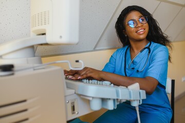 Young female African-american doctor working on modern ultrasound equipment. Operator of ultrasound scanning machine sitting and looking at the monitor, waiting for patient.