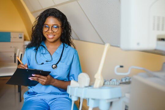 Young african-american female doctor in white coat using ultra ultrasound scanning machine and looking on the screen. African woman doctor working on modern ultrasound equipment.