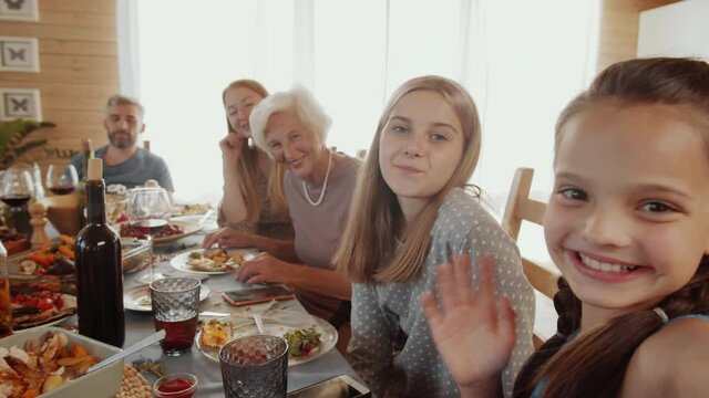 Beautiful little girls, grandmother and cheerful parents looking at camera, smiling and waving while taking selfie together at family dinner