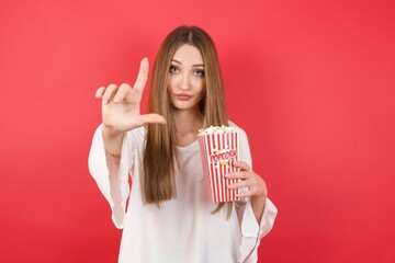 Eastern European woman holding bucket with popcorn standing over isolated red background making fun of people with fingers on forehead doing loser gesture mocking and insulting.