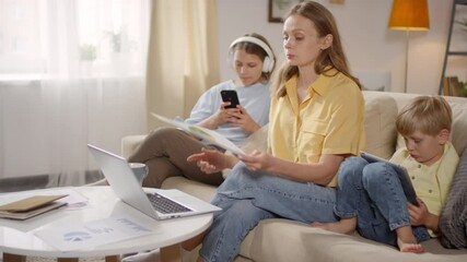 Handheld shot of woman sitting on couch in living room and discussing financial document with colleagues via video call. Little boy with tablet and teenage girl with mobile phone sitting beside her - Powered by Adobe