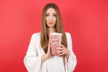 Positive Eastern European woman holding bucket with popcorn standing over isolated red wall smiles happily, glad to receive pleasant news from interlocutor, keeps palms together, People emotions