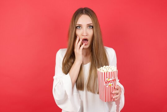 Scared terrified Eastern European woman holding bucket with popcorn standing over isolated red wall shocked with prices at shop, being short of money to buy something, People and human emotions