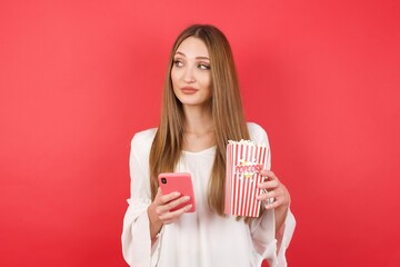 Eastern European woman holding bucket with popcorn standing over isolated red background holds telephone hands reads good youth news look empty space advert