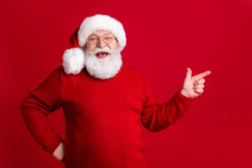 Fototapeta na wymiar Photo of positive old man in santa claus headwear point index finger copyspace indicate jolly holly fairy x-mas magic adverts wear jumper isolated bright shine color background