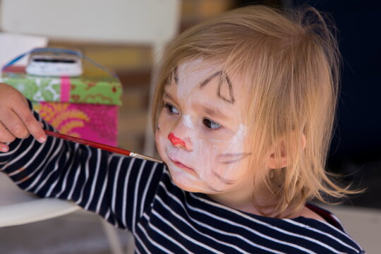 Side closeup of cute blonde toddler girl with serious expression holding a brush to her face with cat painting, Quebec City, Quebec, Canada