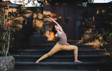 Fototapeta na wymiar Positive caucasian female in tracksuit stretching muscles standing in asana on practice during vacation retreat, calm concentrated woman enjoying relaxation and vitality on yoga training near temple