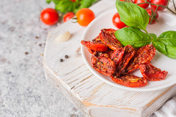 Sundried tomatoes on the white plate on a white wooden board on a gray background. Next to the fresh tomatoes and green basal. Close-up, space for text, copy space