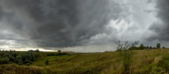Summer panoramic landscape with ominous clouds in overcast sky over the valley wih hills and fields.