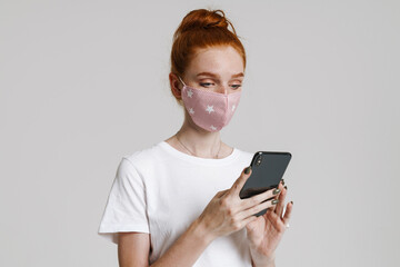 Fototapeta premium Image of focused ginger girl in face mask using mobile phone