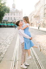 Fototapeta premium Adorable couple in love, enjoying their walk on a sunny day in the city, standing on tram track. Handsome guy holds his attractive young girldriend in blue dress on hands