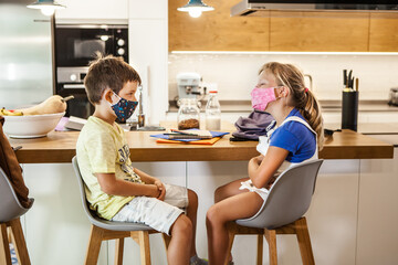 6-years-old caucasian boy and girl with colored protective masks looking at each other. Happy Preschool Kids talking in a house kitchen. Daytime, first day of school after coronavirus pandemic.