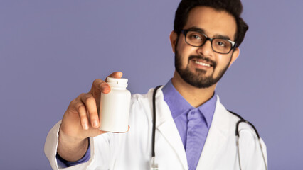 Smiling Indian doctor showing jar of pills on violet background, mockup for design. Selective focus