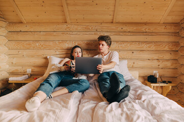 Young beautiful couple lying on bed in wooden apartment and resting, young man showing laptop to woman with book. Leisure at the hotel.