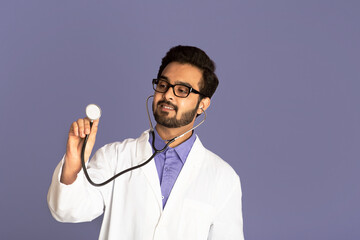 Young Indian doctor in lab coat using stethoscope on lilac background, free space